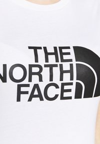 Bílý tričko vyrobené z bavlny, s velkým černým logem, na kterém je v tučných písmenech napsáno "THE NORTH FACE" a ikoničtější zakřivená grafika.