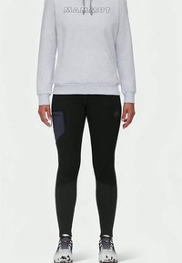 Személy világosszürke Mammut kapucnis pulóvert, fekete leggingset sötét folttal a combján és fehér sportcipőt visel fekete részletekkel.