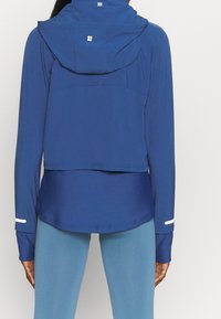 Blå sportjacka med huva, med långa ärmar, en kortad design och vita detaljer på ärmsluten, tillverkad av ett mjukt, lätt material.