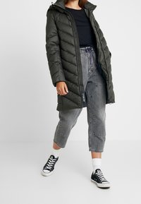 Mörkgrön vadderad jacka med huva, med quiltat mönster, dragkedja framtill och sidofickor, kombinerad med grå korta jeans och svarta sneakers.