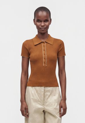 Femme mince portant un polo côtelé marron à manches courtes ajusté et un pantalon beige ample, debout devant un fond clair uni.