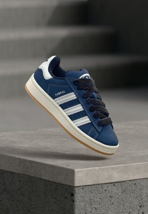 Pantof sport albastru cu dungi albe și șireturi negre, etichetat „CAMPUS”, plutind deasupra unei margini de beton gri, cu scări neclare în fundal.