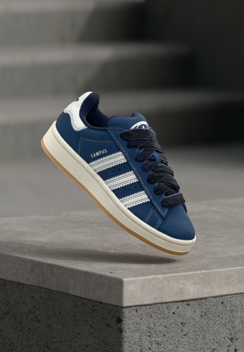 Μπλε αθλητικό παπούτσι με λευκές ρίγες και μαύρα κορδόνια, με την ετικέτα «CAMPUS», αιωρείται πάνω από μια γκρίζα τσιμεντένια ακμή με θολή σκάλες στο φόντο.