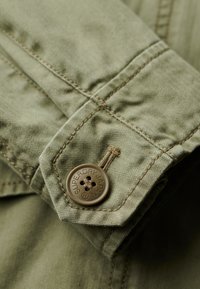 Olijfgroene jasmanchet met een visgraattextuur, voorzien van een bruine knop met de opdruk "SUPERDRY JAPAN" en een gestikte naaddetail.