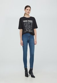 T-shirt grafica nera con la scritta "NEW YORK", abbinata a jeans skinny blu a vita alta e stivaletti neri. Sfondo neutro.