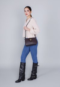 Borsa a tracolla in pelle marrone con fibbia dorata, texture liscia. Indossata con jeans skinny blu e stivali neri fino al ginocchio. Modella in posa di lato.