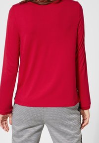 Maglia rossa a maniche lunghe con una texture liscia. Girocollo semplice e vestibilità comoda, abbinata a pantaloni a quadri bianco e nero.