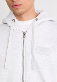 Svetlo šedá mikina na zips s viazačkami, so štruktúrovaným povrchom a značkovou nášivkou na ľavej hrudi. Viditeľný strieborný zips.