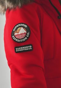 Röd parkas med en foder av päls i huvan, två distinkta runda märken: ett med bergsgrafik och det andra med varumärkesnamnet i vit text.