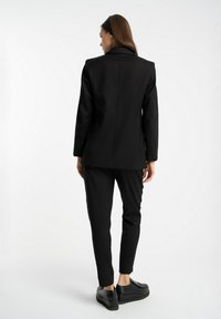 Czarna marynarka dopasowana z wyraźnymi ramionami, w zestawieniu z dopasowanymi czarnymi spodniami. Gładki materiał z minimalnymi detalami, charakteryzujący się prostą sylwetką.
