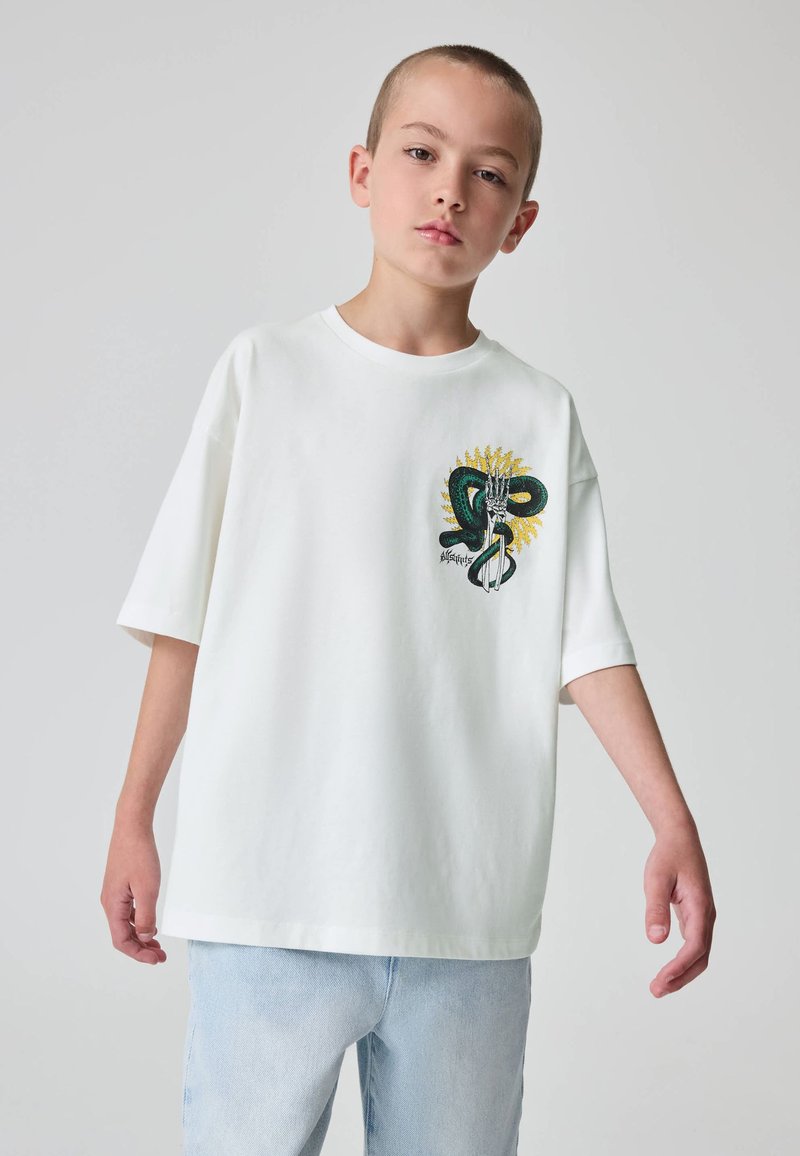 Balti medvilniniai trumpomis rankovėmis marškinėliai su žalia gyvatės piešiniu ir geltonais akcentais priekyje. Laisvas siluetas, minkšta tekstūra.