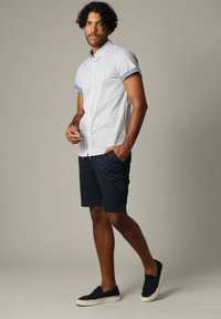 Korte mouwen, geruite blouse in lichtblauw en wit met blauwe manchetten, gecombineerd met marineblauwe shorts en marineblauwe instappers. Neutrale achtergrond.