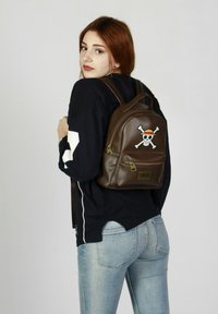 Mochila de cuero marrón con emblema de calavera y tibias cruzadas en blanco, bolsillo con cremallera y correas ajustables, llevada en la espalda de una persona.