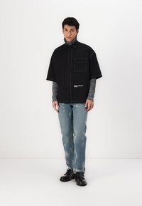 Juodas trumpas rankovės marškinėliai su priekinėmis kišenėmis ir baltomis siūlėmis, dėvimi ant pilkos tekstūruotos golfo, derinami su nusidėvėjusiais šviesiai mėlynais džinsais.