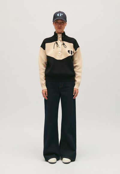 Pull noir et crème à bloc de couleur avec patte de boutonnage et cordons, associé à un pantalon en denim sombre évasé et des baskets blanches.