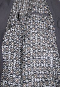 Graue Jackeninnenseite mit einem gemusterten Futter aus geometrischen Formen in Beige, Weiß und Marineblau. Enthält eine kleine Tasche und Knöpfe.