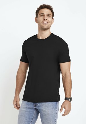 Mann mit kurzen lockigen Haaren lächelnd, trägt ein schwarzes kurzärmliges T-Shirt, hellblaue Jeans und eine schwarze Armbanduhr, steht vor weißem Hintergrund.