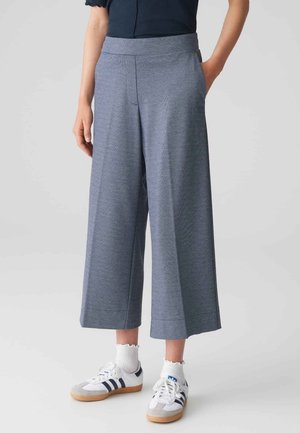 Persona con pantalones anchos grises recortados, calcetines blancos con bordes con volantes y zapatillas blancas con rayas negras.
