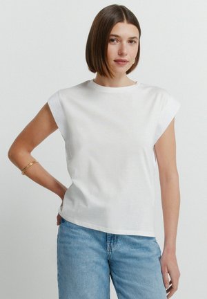 Kobieta o prostych brązowych włosach nosi biały bezrękawnik i niebieskie dżinsy, stoi na tle jednolitego jasnego tła z jedną ręką na biodrze.