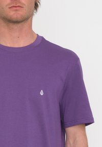 Camiseta de algodón púrpura con mangas cortas, escote redondo y un acento de logo blanco en el lado izquierdo del pecho. Textura suave y ajuste casual.