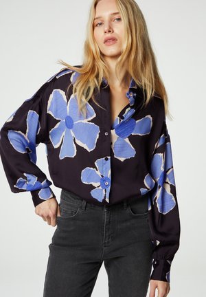 Overmaat zwart shirt met groot blauw bloemenpatroon, lange mouwen en knoopsluiting. Gecombineerd met donkergrijze jeans. Glad stoftextuur.