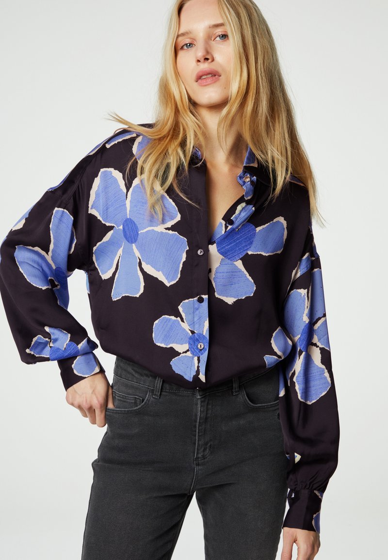 Overmaat zwart shirt met groot blauw bloemenpatroon, lange mouwen en knoopsluiting. Gecombineerd met donkergrijze jeans. Glad stoftextuur.