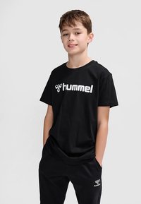 Svart bomulls-t-shirt med korta ärmar och rund hals, med en stor vit "hummel"-logotyp på framsidan. Kombinerad med svarta byxor.