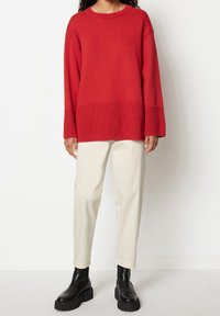Pull rouge en maille avec une texture côtelée, manches larges et bande unie en bas, associé à un pantalon beige clair et des bottines noires.