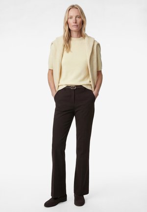 Frau mit langen blonden Haaren steht mit Händen in den Taschen, trägt ein cremefarbenes Kurzarmoberteil, einen passenden Pullover über den Schultern, schwarze Hose und dunkle Schuhe.