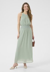 Mint green maxi dress with halter neckline, pleated fabric, lace detailing at the waist, and sheer overlay. Paired with beige heeled mules.
