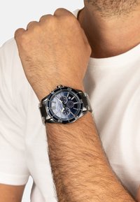 Męski zegarek ze stalową bransoletą, niebieską i srebrną tarczą, trzema subtarczami i cyframi rzymskimi; noszony na nadgarstku o jasnej karnacji.