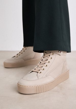 Baskets montantes beige clair avec semelles épaisses côtelées portées avec un pantalon foncé sur un fond de sol clair.