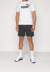 Camiseta blanca con el logo negro de "PUMA", pantalones cortos negros, zapatillas deportivas blancas con acentos rojos y calcetines blancos con un logo negro.