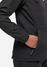 Veste softshell noire avec une poche zippée et des poignets élastiques. Texture lisse avec une coupe ajustée et un ourlet légèrement arrondi.