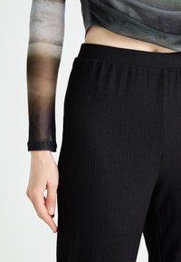 Pantalons noirs texturés avec une taille froncée, associés à un haut transparent à manches longues arborant un motif dégradé dans des couleurs atténuées.
