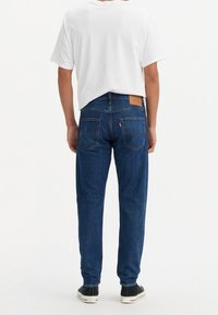 Personne portant un jean bleu, un t-shirt blanc et des baskets noires, debout de dos sur un fond blanc uni.
