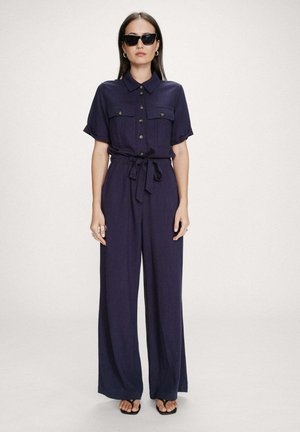 Femme portant une combinaison bleu marine à boutons avec manches courtes, taille nouée, jambes larges, lunettes de soleil noires et sandales noires à lanières, debout devant un fond uni.