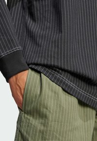 Svart långärmad skjorta med vita vertikala smala ränder, ihop med olivgröna byxor med liknande randigt mönster; båda har kontrasterande detaljer.