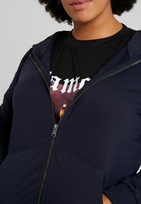 Námořnická zipová mikina s hladkým povrchem, se bočními kapsami a žebrovanými manžetami, navrstvená přes černé tričko s grafikou a tučným bílým textem.