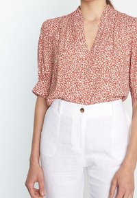 Blusa rossa a fiori con maniche corte e scollatura a V, abbinata a pantaloni in lino bianchi con un bottone e tasche frontali.