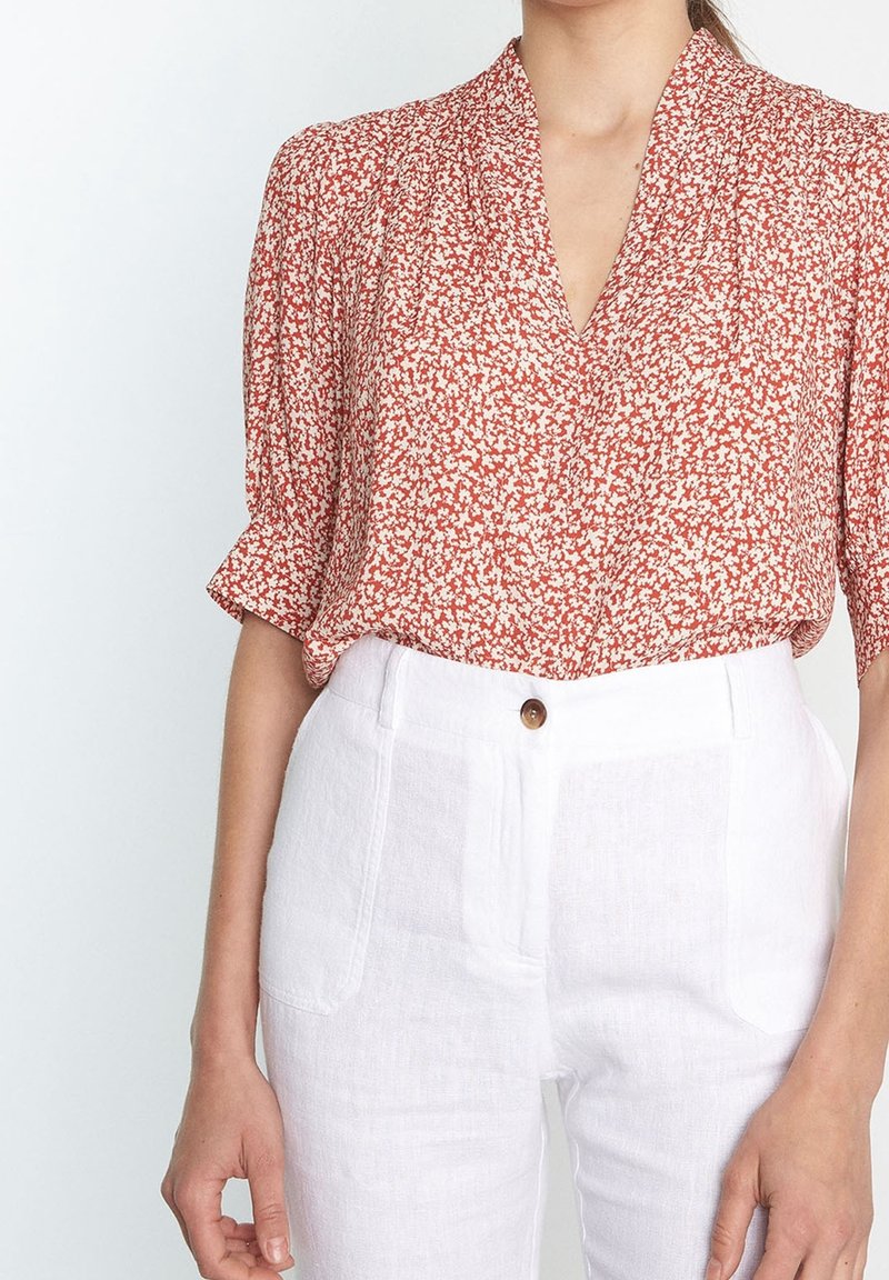 Blusa rossa a fiori con maniche corte e scollatura a V, abbinata a pantaloni in lino bianchi con un bottone e tasche frontali.
