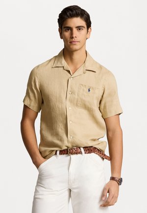 Camisa de lino beige de manga corta con un bolsillo en el pecho, que presenta un logotipo sutil. Llevada con pantalones blancos y un cinturón trenzado marrón.