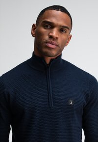 Marineblauwe zip-up trui met een gestructureerd patroon, hoge kraag en een klein zwart logo-opdruk op de borst. Gladde stofafwerking.