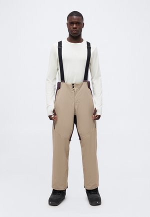 Hombre vestido con pantalones de nieve beige con tirantes negros, camisa blanca de manga larga y botas negras sobre un fondo blanco liso.