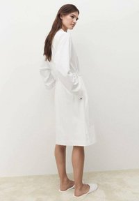 Rochie din bumbac alb, cu mâneci lungi, talie strânsă și buzunare laterale; textură netedă cu o lungime până la genunchi; design simplu.