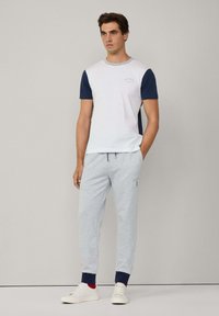Camiseta blanca con mangas azul marino y ribete gris, combinada con pantalones jogger gris claro con puños azul marino y un pequeño logo. Zapatillas blancas completan el atuendo.