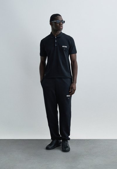 Άνδρας που φοράει μαύρο πόλο πουκάμισο Moschino, ταιριαστό παντελόνι, μαύρα αθλητικά παπούτσια και σκούρα γυαλιά ηλίου, στέκεται μπροστά σε ανοιχτό γκρι φόντο.