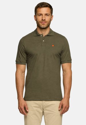 Uomo che indossa una polo verde oliva a maniche corte e pantaloni beige, in piedi davanti a uno sfondo chiaro e semplice.
