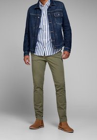 Giacca di jeans, camicia a righe con bottoni, pantaloni verde oliva e stivali color nocciola. Caratteristiche includono un taglio classico e colori a contrasto.
