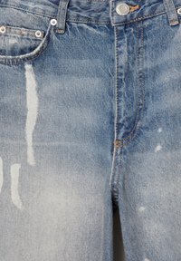 Jean en denim bleu avec des zones délavées et usées, présentant des coutures visibles, des poches avant et une fermeture par bouton en métal à la taille.